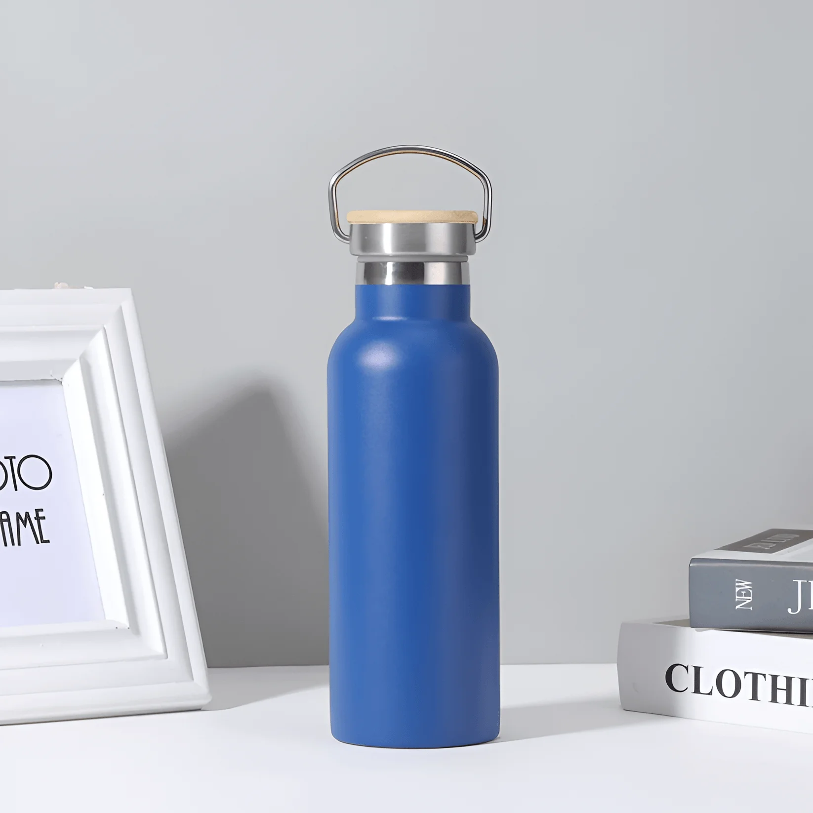 Minimalist insulated stainless steel water bottle with handle lid, reusable thermos bottle on a clean desk setup with books and picture frame in background.