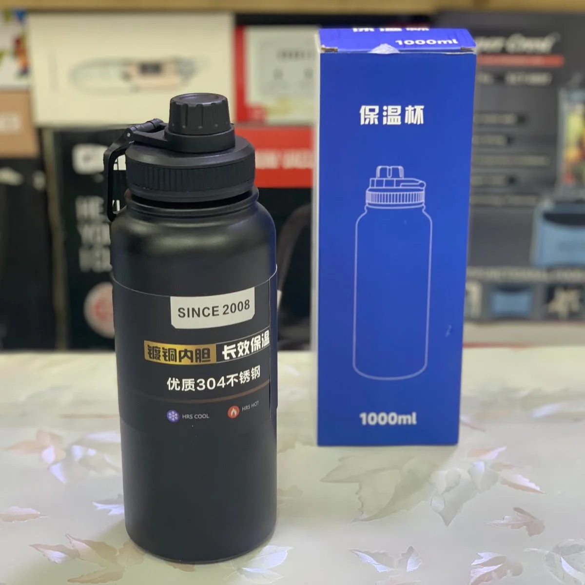 A black 1000ml insulated stainless steel vacuum water bottle with a carry handle and screw cap, placed beside its blue packaging box featuring Chinese text and a line illustration of the bottle.